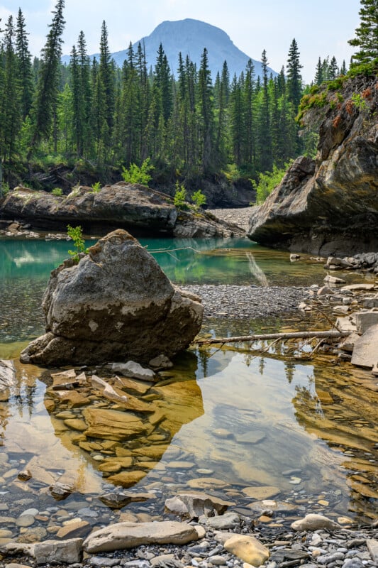 A serene mountainous landscape with a foreground of clear, shallow water revealing smooth rocks underneath. A large boulder stands in the water, and tall pine trees line the shore. In the background, a majestic mountain peak rises under a partly cloudy sky.