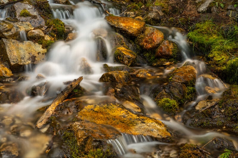 A serene forest stream flows over and around moss-covered rocks and logs, creating cascading waterfalls. The water appears silky smooth due to long exposure photography, enhancing the tranquil atmosphere of the natural scene.