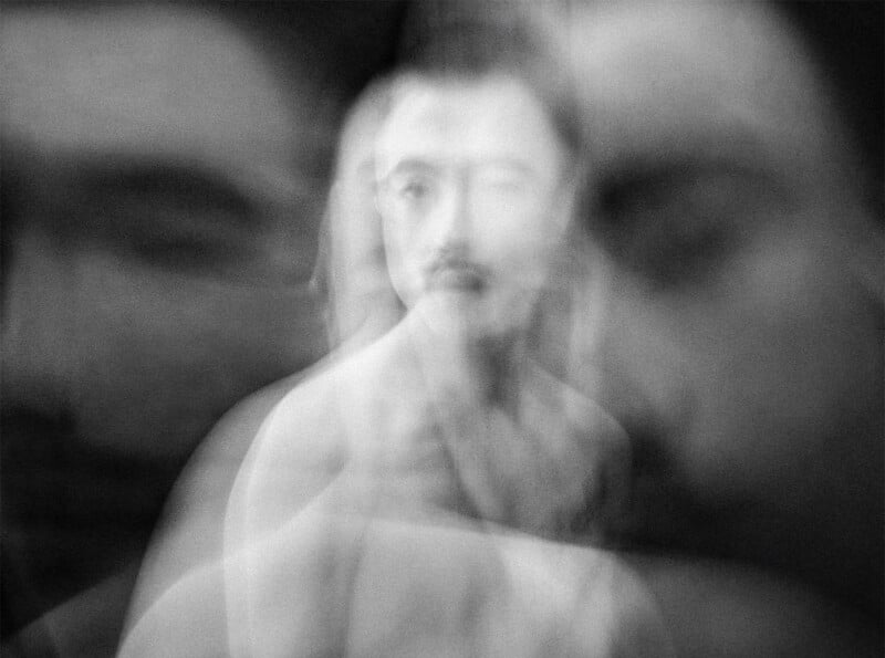 A monochromatic, blurry image showcasing multiple overlapping faces of a person with a mustache. The figures blend and blur together creating a ghostly, ethereal effect. The overall mood is abstract and surreal.