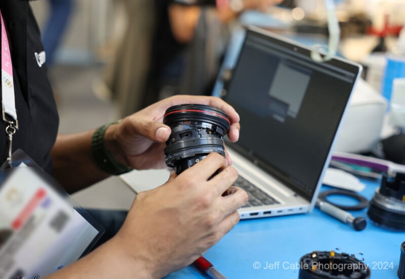 A person is holding a camera lens and performing maintenance or repair work on it. There is a laptop in the background with a blurred screen, along with some tools and equipment on the table. The photo is credited to Jeff Cable Photography 2024.