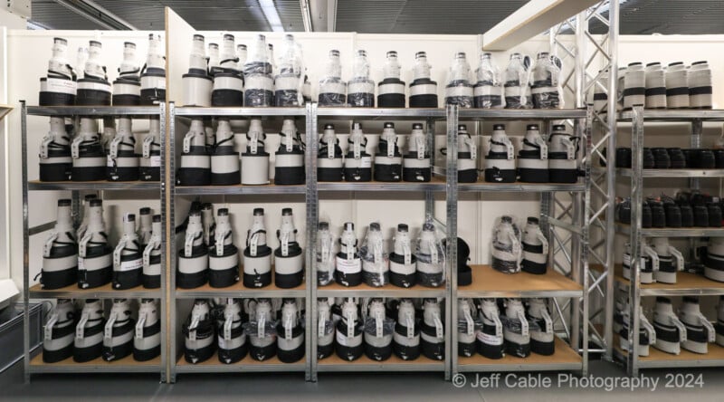 A storage room filled with multiple shelves containing large camera lenses and other photography equipment. The lenses are organized and neatly placed in rows. The photo is credited to "Jeff Cable Photography 2024.