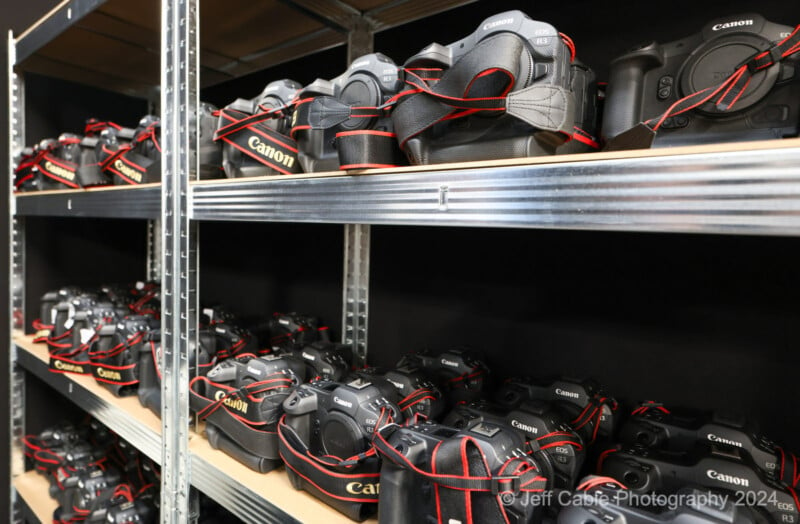 A metal shelving unit holds multiple rows of Canon DSLR cameras with red and black straps. Each shelf has several cameras neatly arranged in a uniform manner. The image is credited to Jeff Cable Photography 2024 in the bottom right corner.