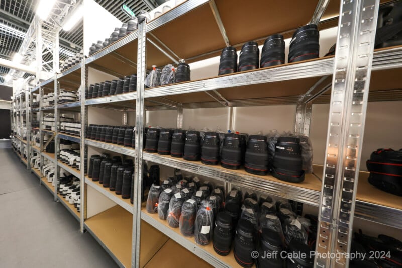 The image depicts metal shelving units lined with numerous camera lenses. The lenses are neatly organized, some wrapped in protective plastic. The background includes additional shelves filled with more camera equipment. Photo credit is given to Jeff Cable Photography 2024.