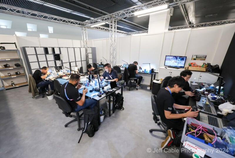 A room filled with people working at different workstations equipped with electronic devices and tools. Some are focused on laptops while others solder components. Storage shelves line the back wall, and a television monitor displays images. © Jeff Cable Photography 2024.