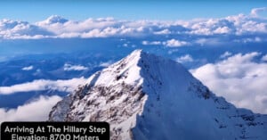 A snow-covered mountain peak rises sharply against a blue sky filled with scattered clouds. The caption reads, "Arriving at The Hillary Step - Elevation: 8700 Meters." The mountain is part of a rugged, vast landscape with distant mountain ranges visible beyond.