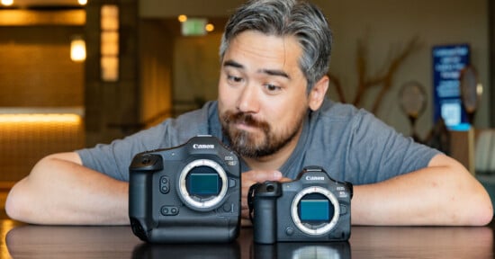 A man with a gray shirt and gray hair and beard curiously looks at two Canon cameras on a table. The left camera is significantly larger than the one on the right. The background is indoors with blurred details and warm lighting.