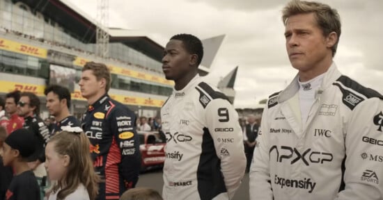 Group of race car drivers standing side by side in racing suits, facing forward. The setting appears to be a racing event with a crowd and track infrastructure in the background. .