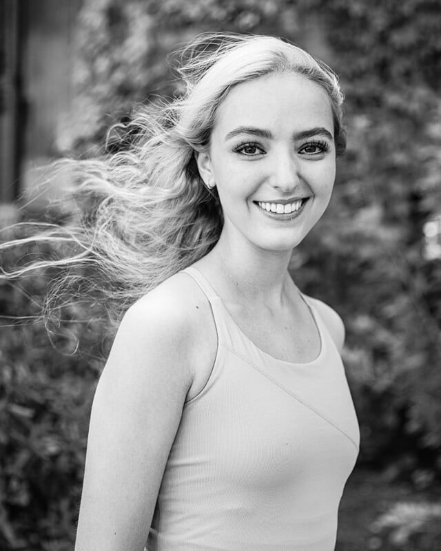 Black-and-white photo of a smiling individual with long, wavy hair blowing in the wind. The person is wearing a sleeveless top and stands against a blurred outdoor background of foliage.