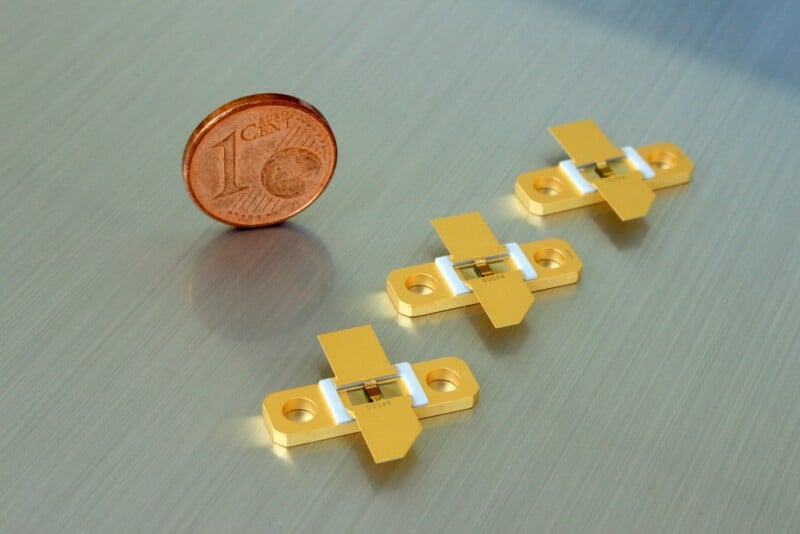 A close-up of a 1-cent euro coin next to three small electronic components with yellow, rectangular bodies and white bases. The components have two holes for mounting and are positioned on a reflective surface.