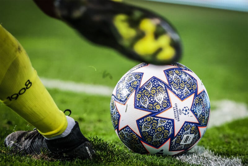 A close-up of a soccer player's foot, wearing a yellow sock and black cleat, about to kick a UEFA Champions League soccer ball adorned with blue and yellow patterns. The ball is positioned on a grassy field near a white line.