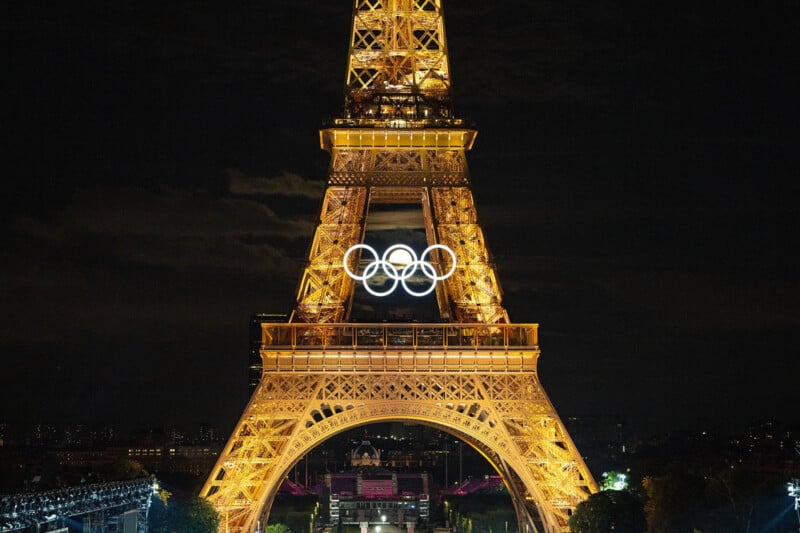 The Eiffel Tower illuminated at night, showcasing the Olympic rings in bright white light hanging prominently at its center. The archway at the base is visible with dark surroundings, highlighting the grandeur of the tower against the night sky.