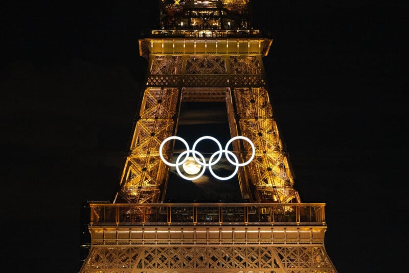 The Eiffel Tower illuminated at night, displaying the Olympic rings in bright lights at its center, signifying the upcoming Paris 2024 Olympics. The metallic structure is bathed in golden light, creating a stunning visual against the dark sky.