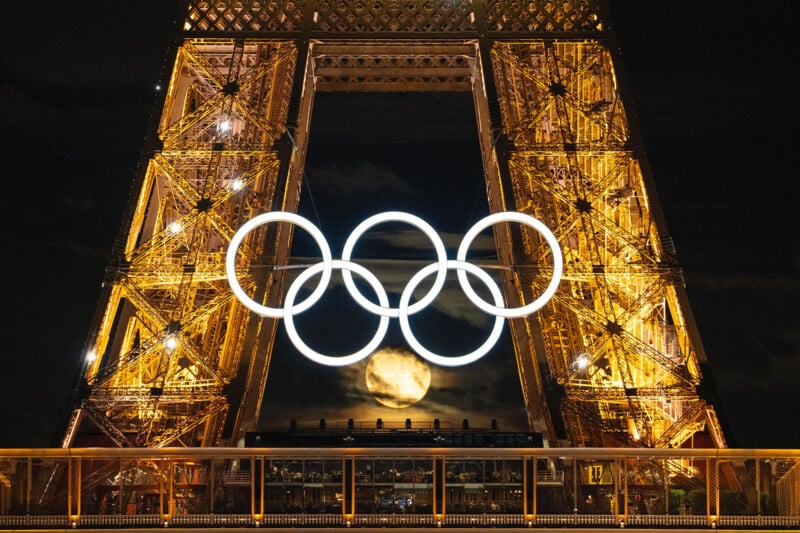 A nighttime view of the Eiffel Tower illuminated with golden lights features large Olympic rings in the center. A bright full moon is visible in the background, framed perfectly between the iron beams of the tower.