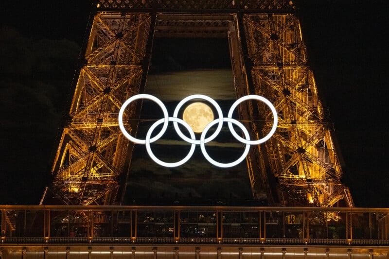 The Eiffel Tower illuminated at night, showcasing the Olympic rings in the center. The full moon glows prominently through the rings, creating a striking visual effect. The metallic structure of the tower glistens with golden lights against the dark sky.