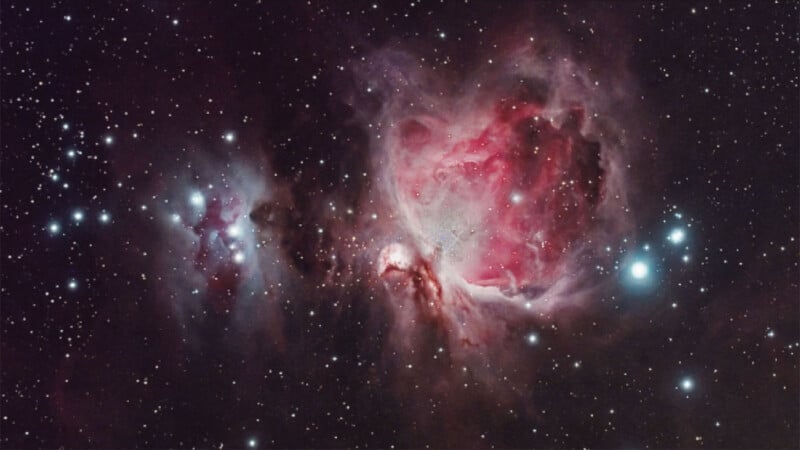 A vibrant image of the Orion Nebula, showcasing a dense region of colorful gas and dust with prominent pink and red hues at its core, surrounded by bright stars scattered across a dark, starry background.