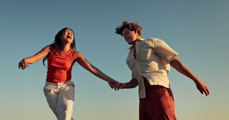 A joyful couple holding hands while laughing and leaping against a clear blue sky. The woman wears a red sleeveless top and white pants, and the man wears a light, short-sleeved shirt and red trousers. They are mid-jump, capturing a moment of carefree happiness.