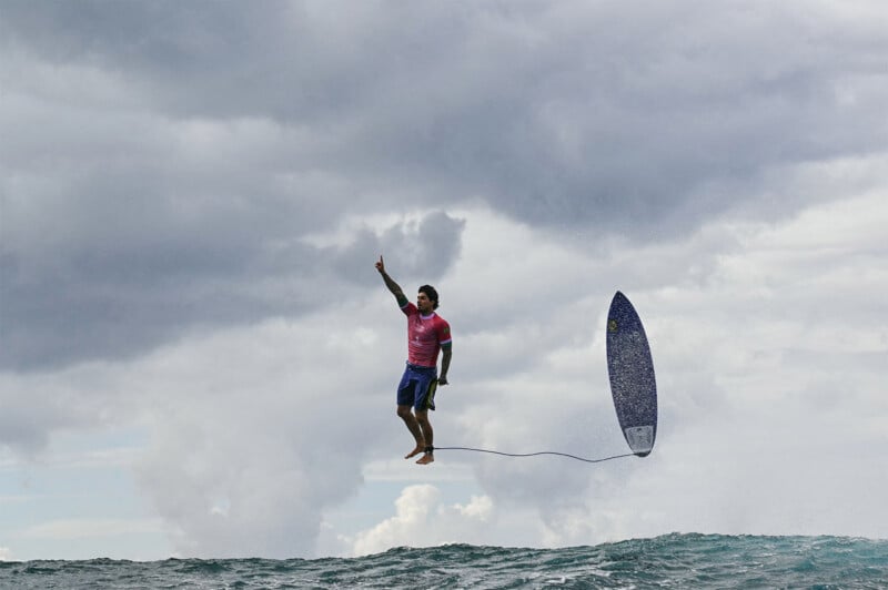 A surfer is captured mid-air while performing a jump and holding a surfboard leash, with the board also airborne beside him. The ocean waves are visible below, and a cloudy sky forms the dramatic backdrop.