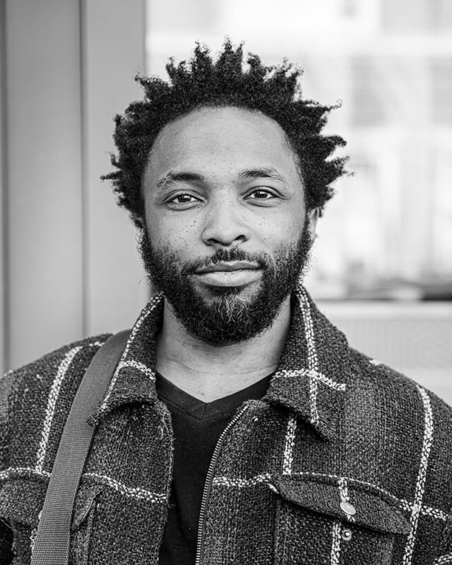 A black and white photo of a man with a short, curly beard and afro-textured hair. He is wearing a checkered jacket over a solid-colored shirt, looking directly at the camera with a slight smile. The background is a blurred urban setting.