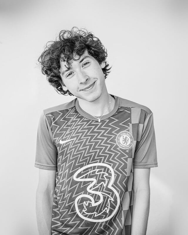 Black and white photo of a young person with curly hair, slightly tilting their head and smiling at the camera. They are wearing a patterned sports jersey with the number three and a logo on the chest. The background is plain and light-colored.