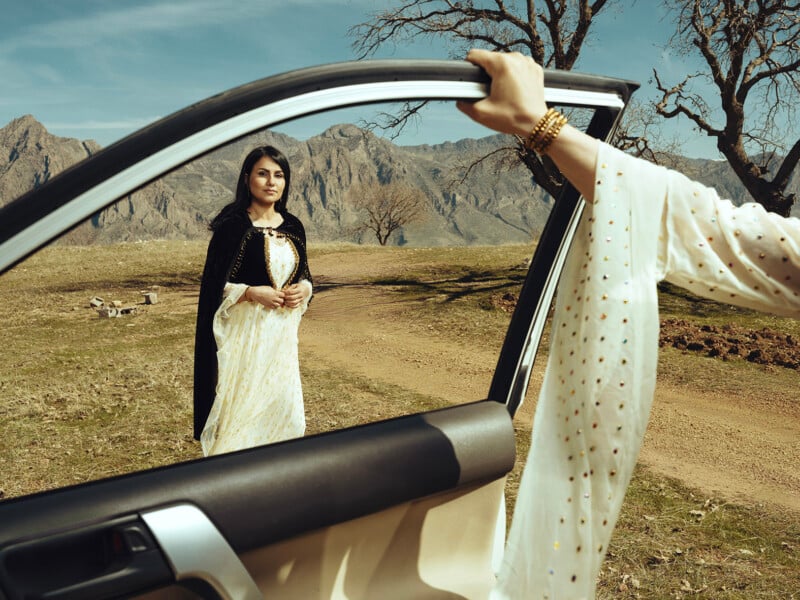 A woman stands outside in a rural area beside a car, looking towards the open driver's side door. She is wearing a white dress with a black cloak. Another woman's hand, dressed in a white, gold-dotted sleeve, is seen holding the car door. Mountains and trees are in the background.