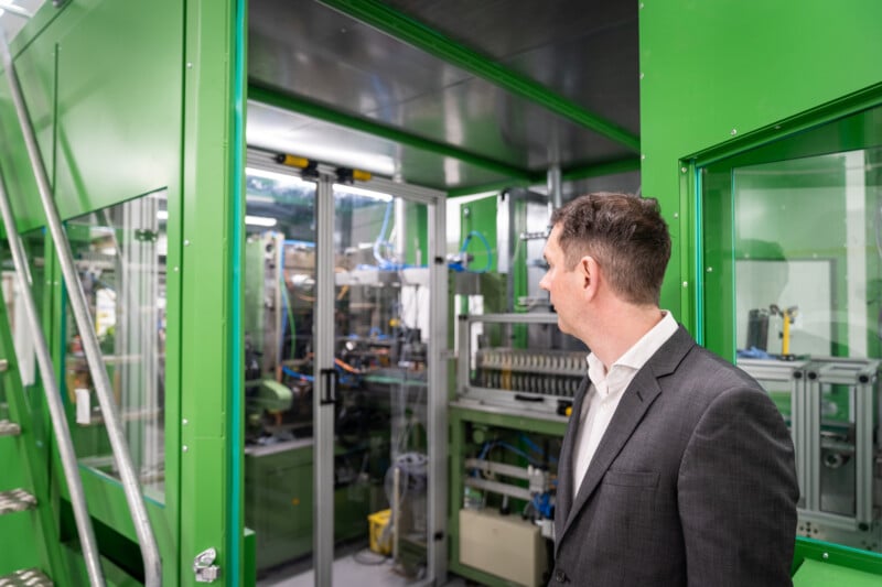 A man in a gray suit observes machinery inside a green, modern industrial facility. The equipment appears complex, with various cables, pipes, and mechanical components visible. The setting is clean and well-lit, suggesting a high-tech manufacturing or production environment.