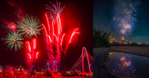 A vibrant fireworks display in a city skyline. On the left, multicolored fireworks illuminate a bridge and buildings with emphasis on red trails. On the right, blue and white fireworks burst against the night sky, reflected on a wet surface below.