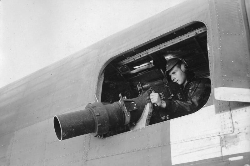 A person in military gear operates a mounted machine gun from an aircraft's side window. The individual is wearing a helmet and leather jacket, focusing intently on the weapon, with the aircraft's metal exterior visible around the window frame.