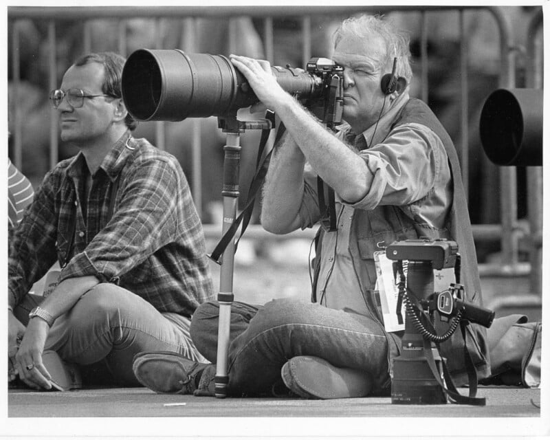 A black and white photo shows an older man seated on the ground using a large telephoto lens on a camera mounted on a tripod. He is wearing headphones and focused on capturing an image. Another man, seated next to him, wears glasses and a plaid shirt.