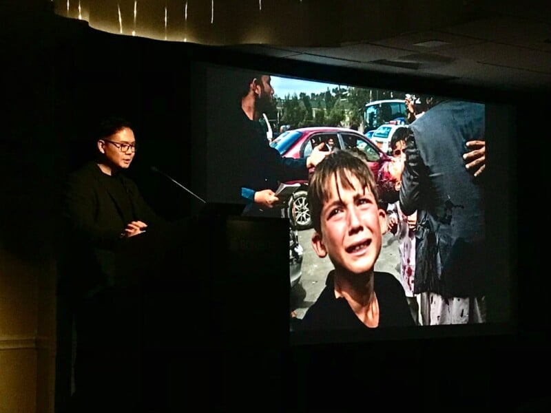 A person stands at a podium presenting a slideshow. The projected image shows a distressed child crying amidst a chaotic street scene with adults around them. The presenter appears to be discussing the photograph in a dimly lit room.