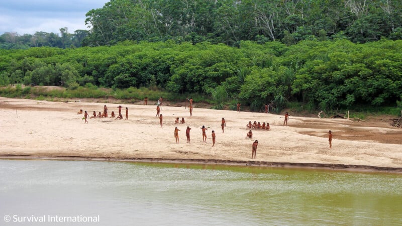 A group of indigenous people, wearing minimal clothing, gathered on a sandy riverbank surrounded by dense green foliage. Some individuals are standing while others are sitting, engaging in various activities. The river flows by the bank under an overcast sky.