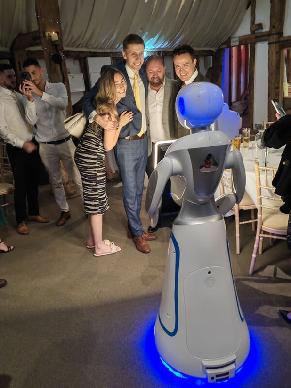 A group of people are gathered around and posing with a humanoid robot in a festive setting. The men are dressed in suits while a woman in a striped dress leans into the group. The scene appears lively with a mix of standing tables and chairs in the background.