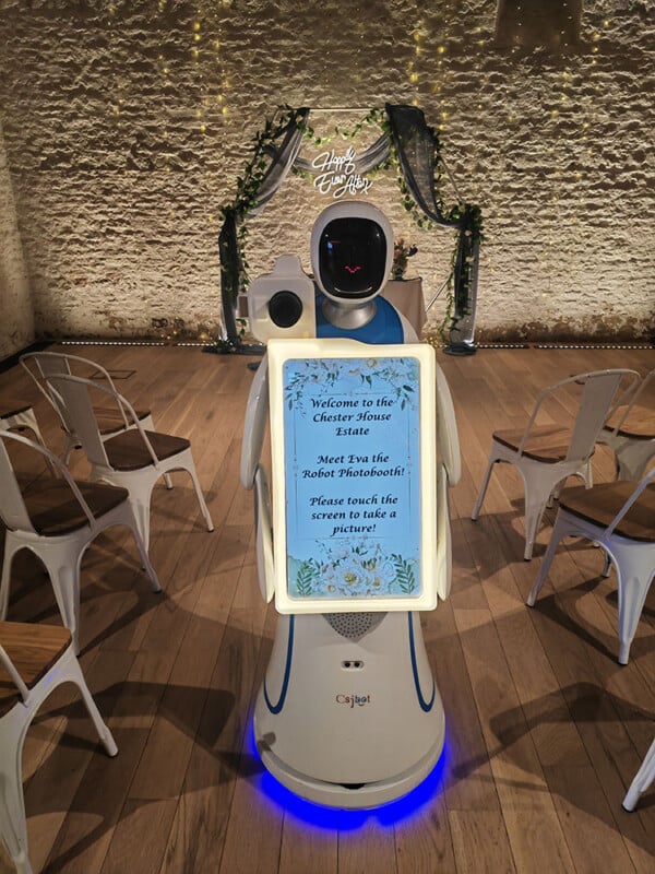 A robot with a rectangular screen stands in a room with wooden floors and white walls. The screen displays a welcome message and instructions for taking a picture. Chairs are arranged in rows, and a wedding arch is decorated with flowers in the background.