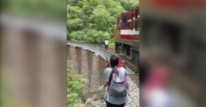 Two people carrying backpacks walk along a narrow train track beside a moving red and white train. The track is elevated with a steep drop on one side, surrounded by dense green foliage. Another person is walking ahead of them on the tracks.