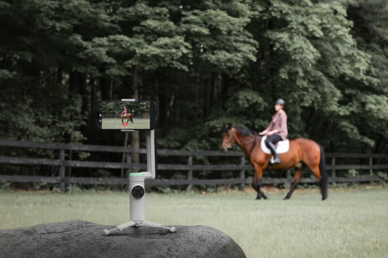 A smartphone mounted on a gimbal tripod focuses on recording a person riding a horse. The rider, wearing a helmet and plaid shirt, guides the brown horse near a wooden fence with dense green trees in the background. The setup is placed on a large rock.