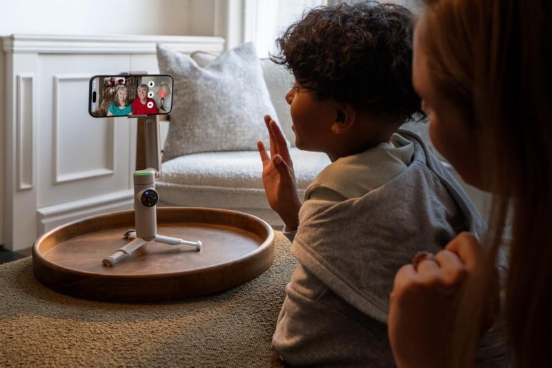 A child and an adult are making a video call using a smartphone mounted on a small tripod. They are seated on a couch, and the phone displays two people on the screen. The child is making a peace sign. The setting is a cozy living room with natural light.
