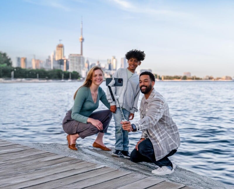 A man, woman, and a young boy take a selfie using a smartphone mounted on a selfie stick. They are on a wooden boardwalk beside a large body of water, with a city skyline, including a tall tower, visible in the background. All three are smiling.