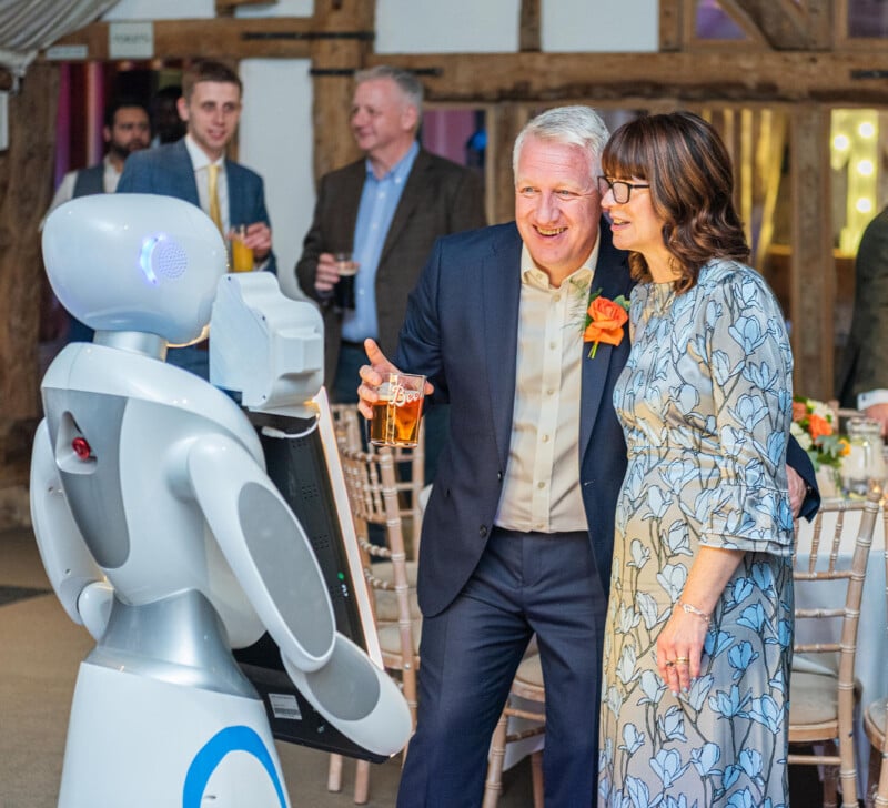 A man and woman smile and pose for a photo with a humanoid robot at an indoor event. The man holds a pint glass, and the woman wears a patterned dress. Other attendees are visible in the background, socializing near wooden beams and bright lighting.