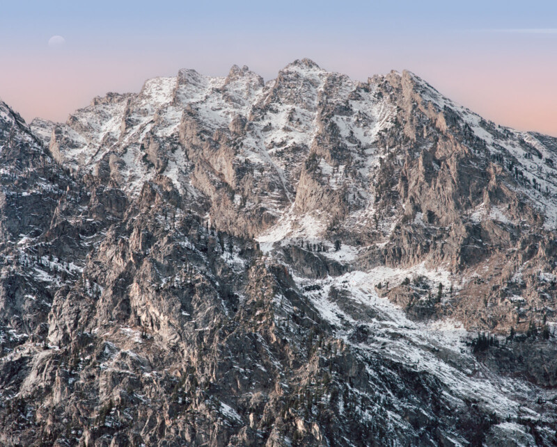 A rugged mountain range with rocky, snow-dusted peaks under a clear sky at dawn or dusk. The moon is faintly visible in the sky, adding to the serene and majestic atmosphere. Sparse patches of vegetation dot the mountainsides.