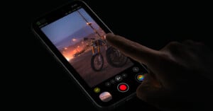 A hand is touching the screen of a smartphone, which displays a photo of a motorcycle under streetlights at dusk. The phone's camera interface is visible, with buttons for settings, shutter, and video recording at the bottom of the screen.