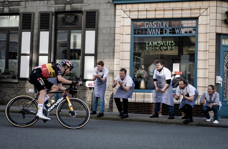 A cyclist dressed in a colorful racing uniform rides past a group of cheering people wearing aprons and blue shirts, who are standing outside a shop labeled "Gaston Van de Walle Lamsvlees Eigen Kweek." The onlookers appear excited and supportive.