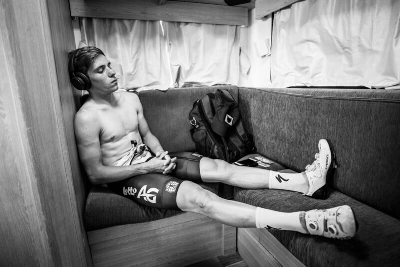A shirtless cyclist with headphones is reclining on a cushioned bench inside a motorhome. He appears relaxed, with his eyes closed and his legs stretched out. He is wearing cycling shorts and shoes, and a backpack is placed next to him. The scene is in black and white.