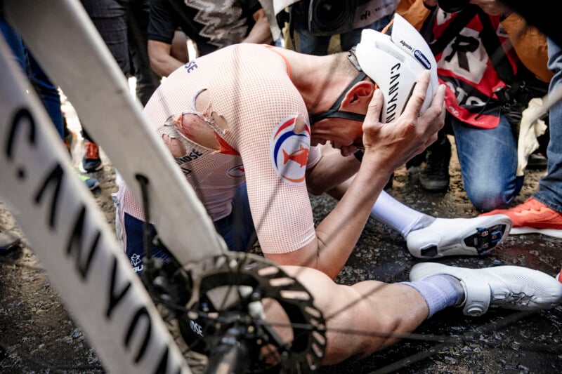 A cyclist wearing a torn jersey sits on the ground, holding his head in distress. Nearby individuals, some with cameras, surround him. A bicycle with the brand name "Canyon" is visible in the foreground. The scene suggests a recent accident or fall.