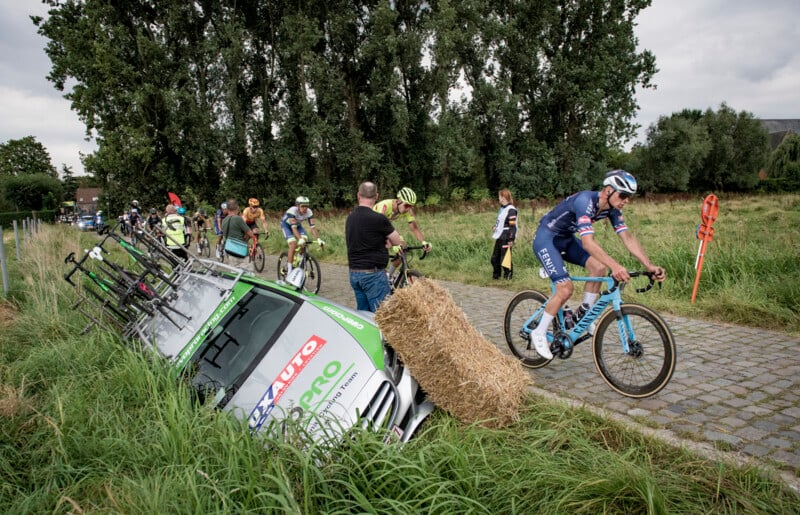Cyclists ride on a cobblestone path while a car, partially off the road and leaning against a hay bale, is on the roadside. People are standing nearby observing the scene. Trees and grassy fields are in the background, under a cloudy sky.