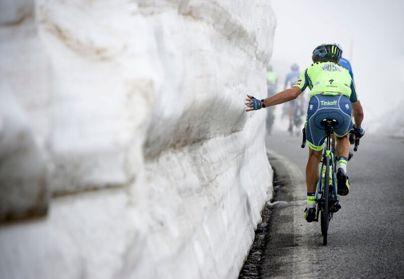 A cyclist wearing a neon green and blue Tinkoff team uniform is riding on a mountain road flanked by high snow walls, touching the snow with one hand. Other riders are visible ahead on the foggy road, creating a dramatic and challenging atmosphere.