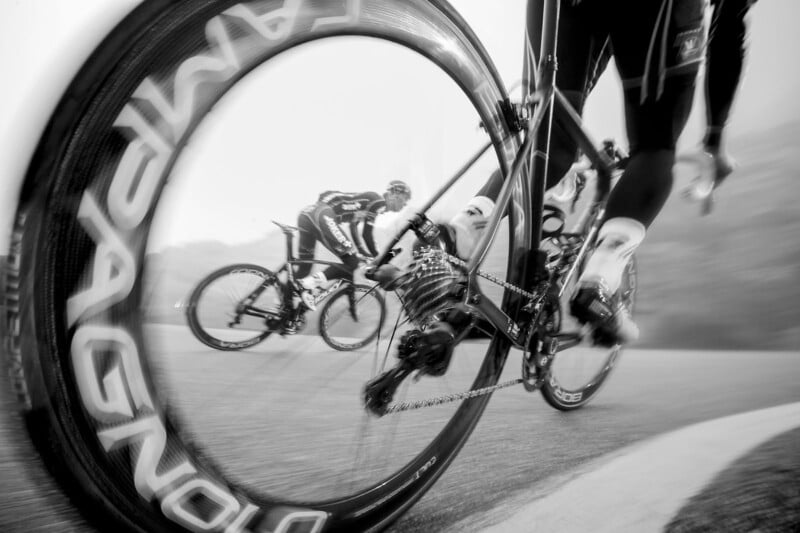 Black and white image of cyclists in a race. The foreground focuses on a close-up of a bicycle wheel with "CAMPAGNOLO" written on it, capturing the motion blur. In the background, two other cyclists are seen racing on a foggy track.