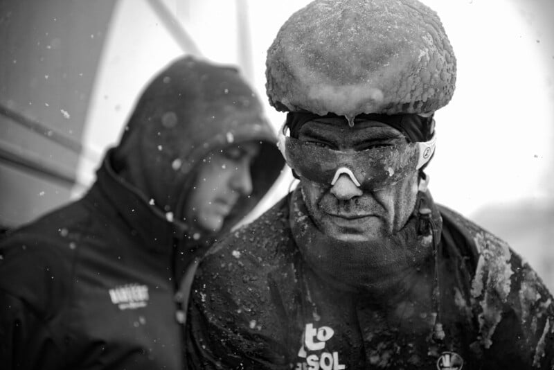 A black-and-white photo shows a person with snow and ice covering their hat, goggles, and jacket, creating a frosty appearance. Another person in the background, also wearing a hooded jacket, is slightly out of focus. Snow is falling around them.