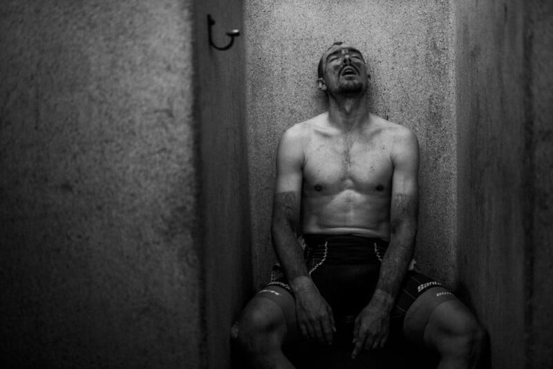 A shirtless man with a sweaty, dirty face and torso sits slumped against a wall in a narrow, dimly-lit space, appearing exhausted. He is wearing athletic shorts and has his head tilted back, eyes closed, and mouth slightly open, suggesting extreme fatigue.