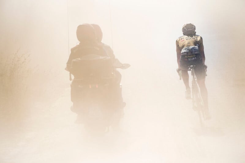 A cyclist is riding on a dusty road, followed closely by two people on a motorcycle. The air is thick with dust, creating a hazy and challenging environment for the cyclist. The scene is backlit, emphasizing the dust and reducing visibility.