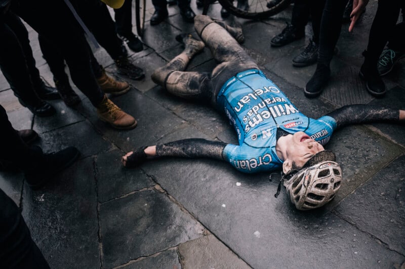 A cyclist in a mud-stained blue jersey lies on their back on a wet pavement, arms outstretched and eyes closed. The helmeted individual is surrounded by onlookers, some with bicycles, suggesting the end of a challenging ride or race.