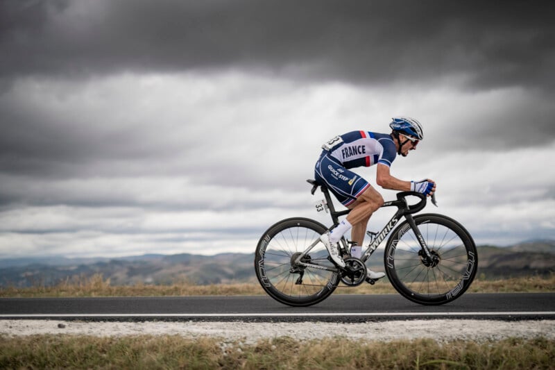 A cyclist in a blue and white racing uniform rides a road bike swiftly along a paved road under a cloudy sky. The background features a hilly landscape. The cyclist's uniform has "France" written on the back.